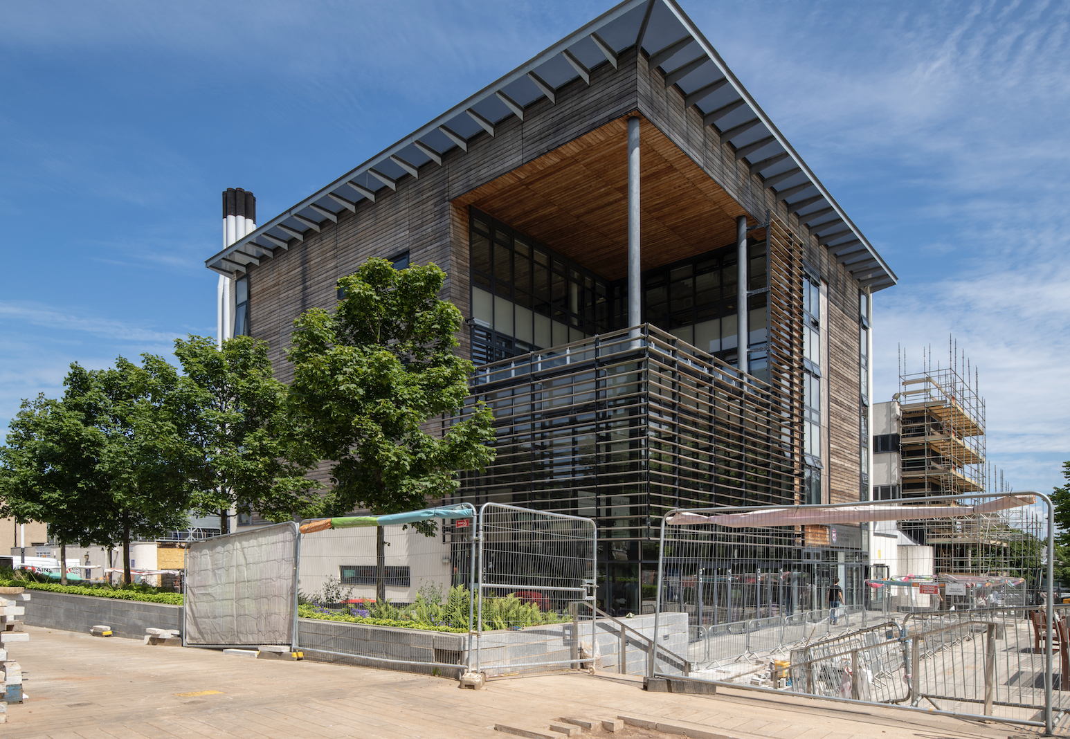 A modern building with wood and glass exterior is under construction, surrounded by scaffolding, metal fencing, and construction materials, with green trees and a clear blue sky in the background.