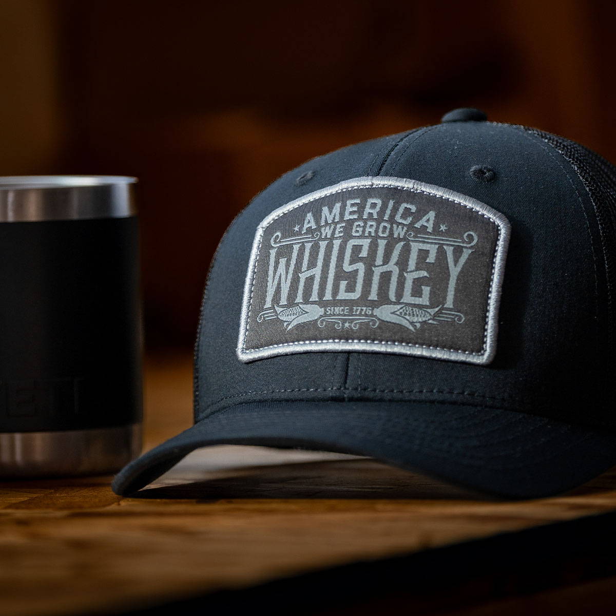 A black hat with a woven patch that says "America We Grow Whiskey". This represents the We Grow Whiskey Collection