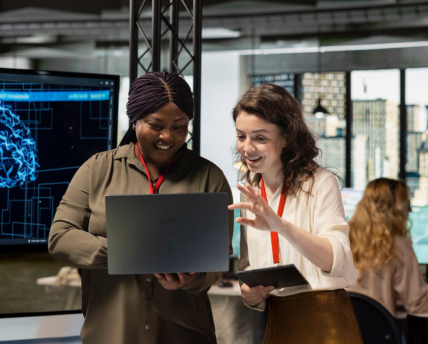 Two women wearing red lanyards smile while looking at a laptop together in a modern office. One holds a tablet, and a digital brain diagram is displayed on a monitor in the background.