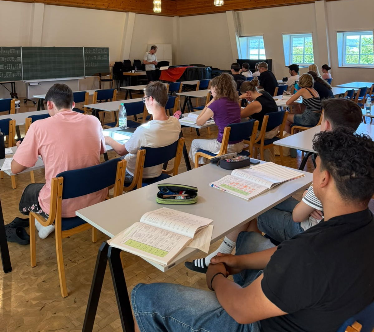 In einem Raum sitzen einige Menschen an Tischen und hören jemandem zu, der vor einer Tafel steht. 