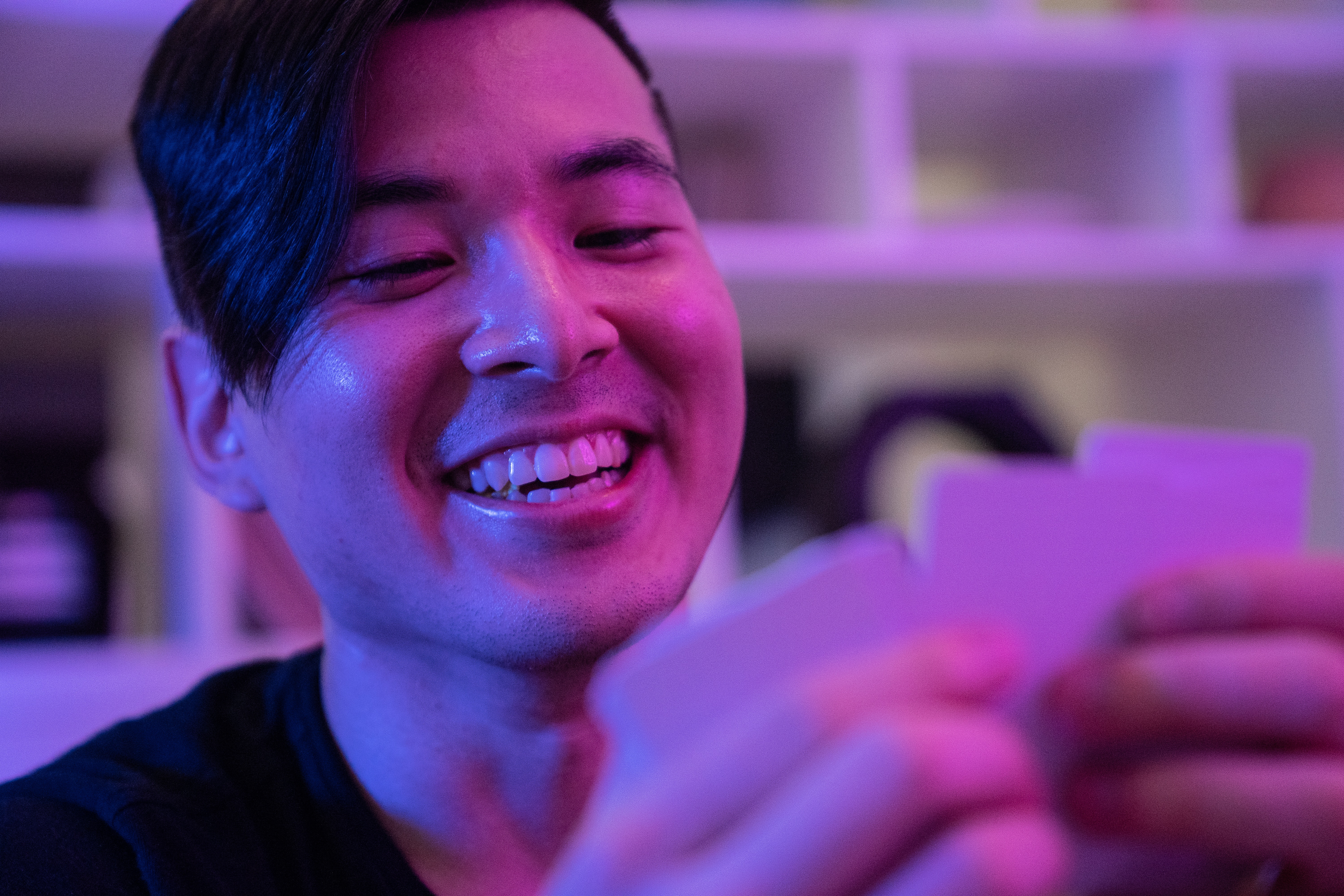 An asian man with long hair smiles as he holds his cards in front of him.