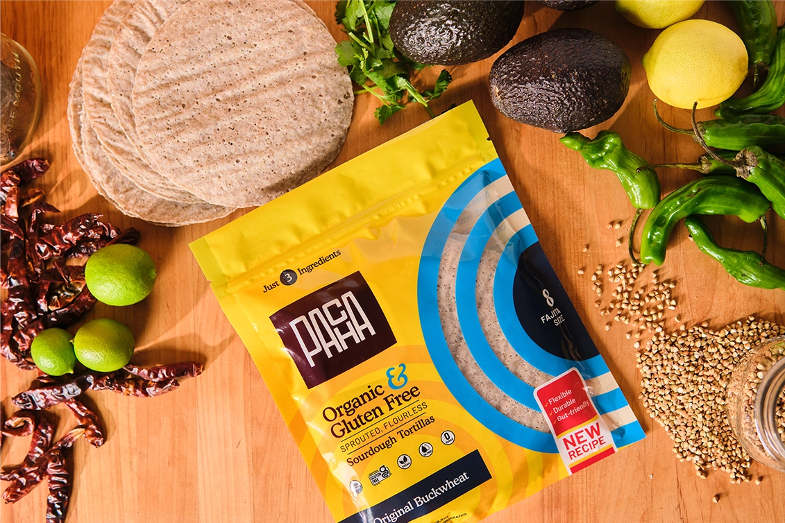 A flat-lay view on a wooden surface featuring a yellow and blue bag of PACHA Organic & Gluten Free Sourdough Tortillas in Original Buckwheat flavor. A stack of grey-brown buckwheat tortillas sits in the top left, surrounded by fresh ingredients including whole avocados, green peppers, lemons, limes, dried red chilis, cilantro, and spilled buckwheat groats.