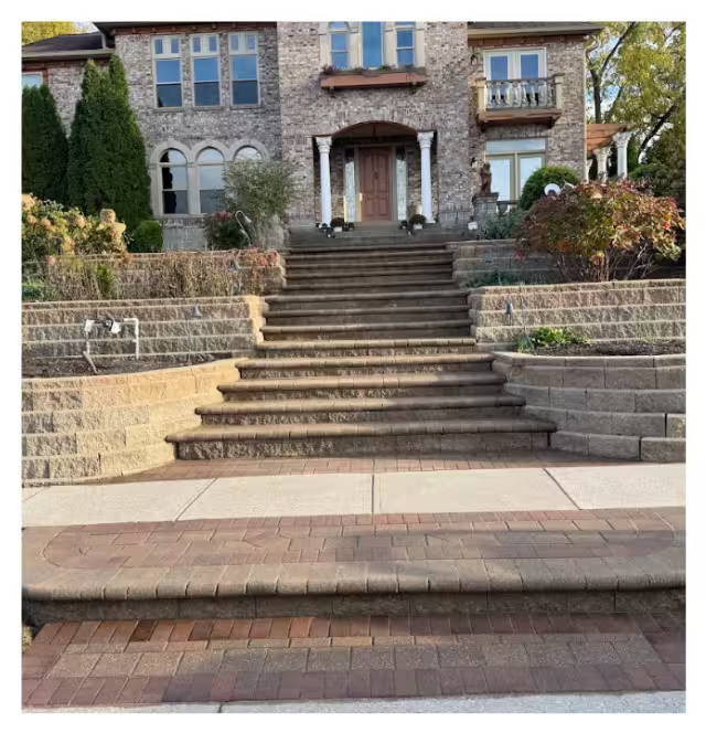 Wide brick and stone staircase with paver landing leading up to a large brick home, bordered by tiered retaining walls and landscaped garden beds.