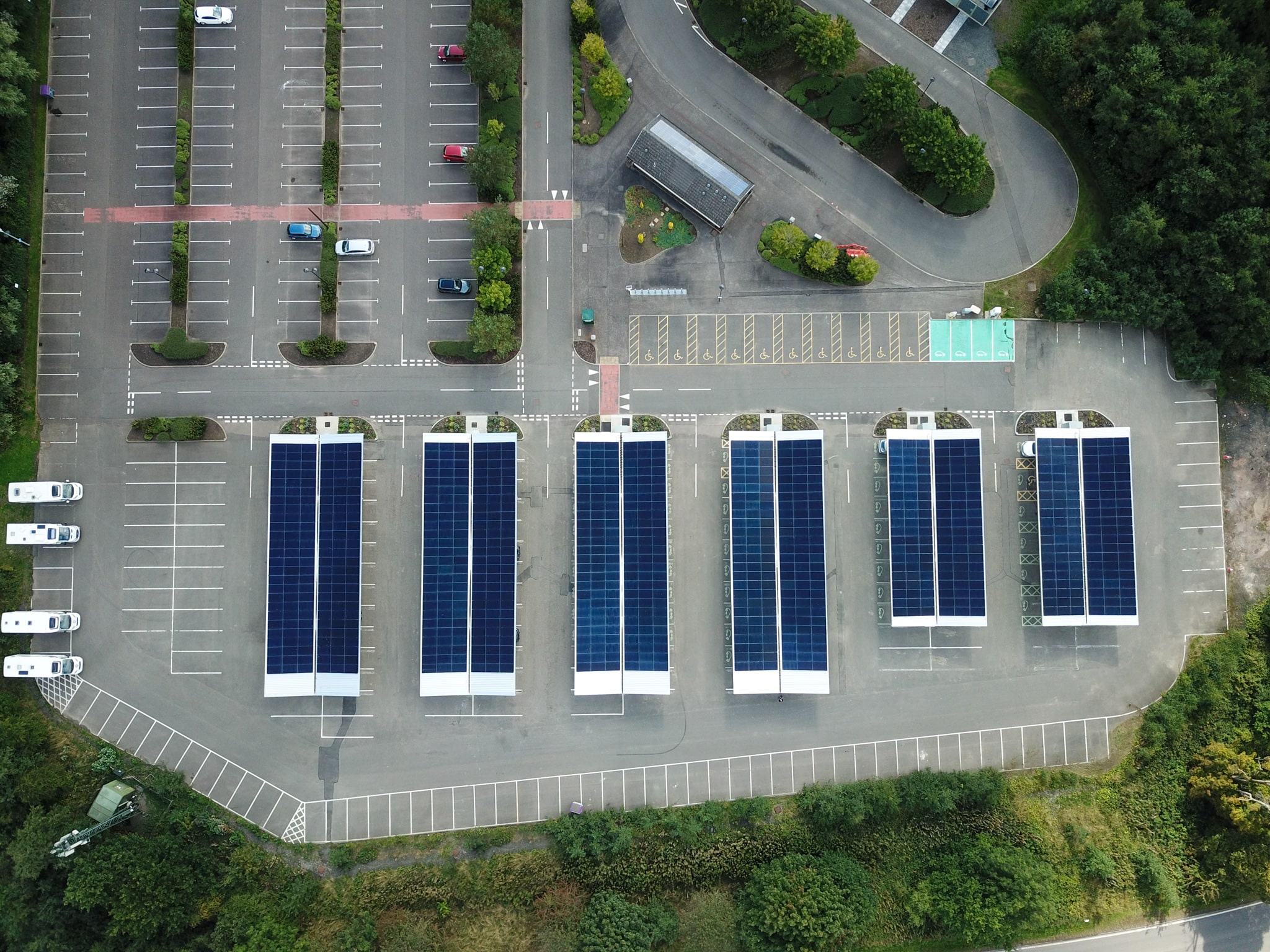 Aerial view of a large parking lot with rows of parking spaces covered by solar panel canopies, surrounded by trees and greenery. Several cars are parked beneath the solar panels.