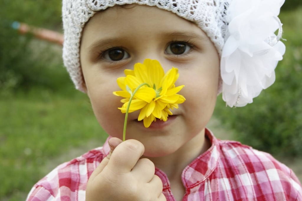 Gros plan sur un jeune enfant portant un bonnet en tricot blanc avec une grande fleur en tissu, tenant une marguerite jaune devant son nez tout en regardant directement l'objectif.