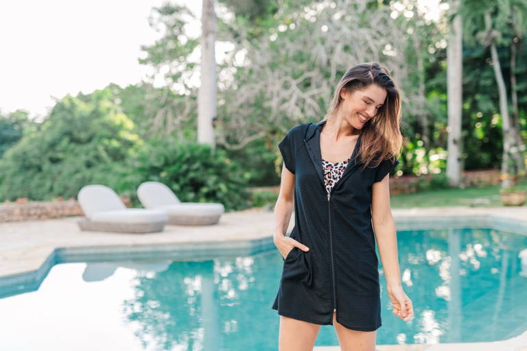 A woman wears a leopard print tankini top and a black cover up while standing by a pool.