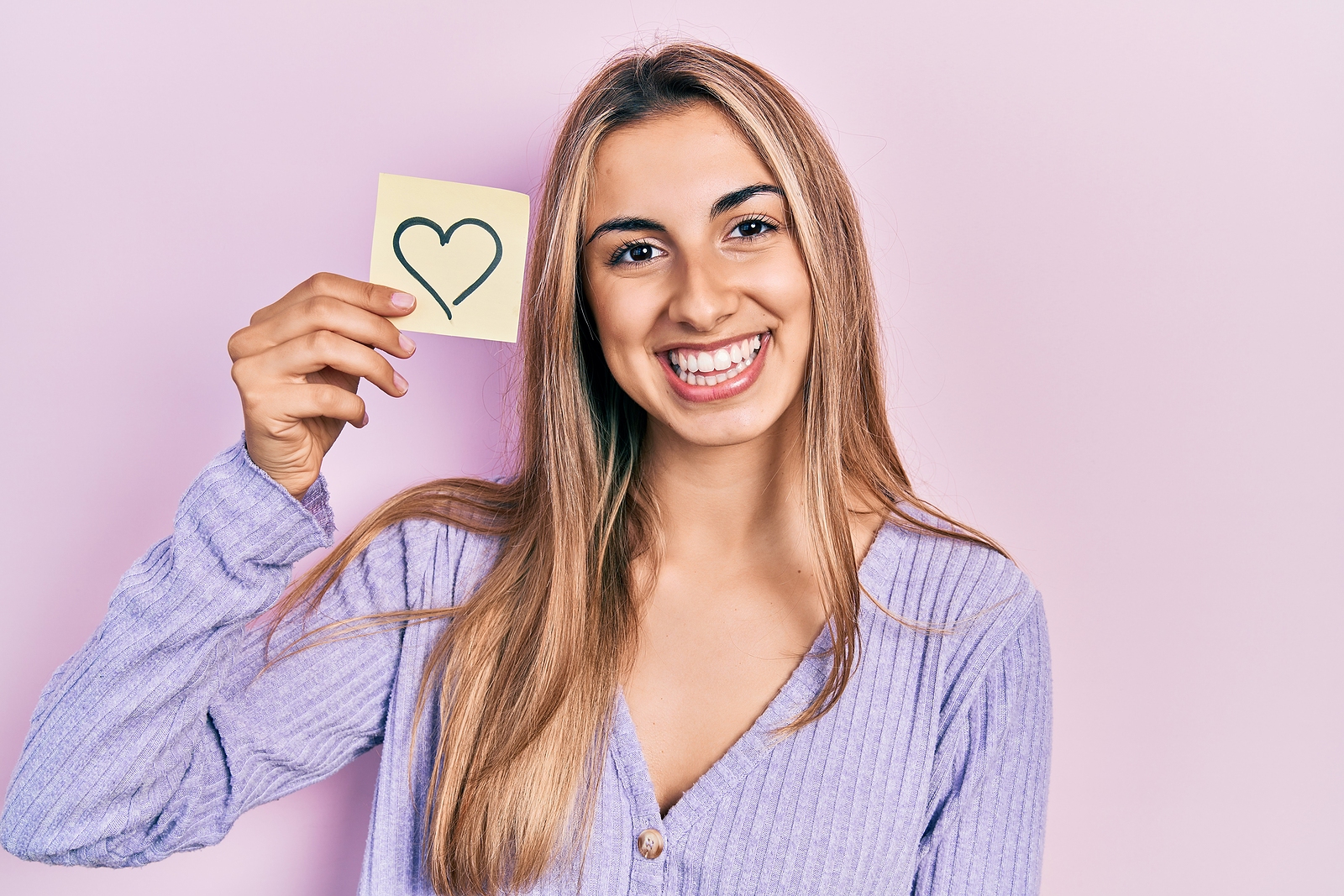 Beautiful hispanic woman holding heart reminder looking positive and happy standing and smiling with a confident smile