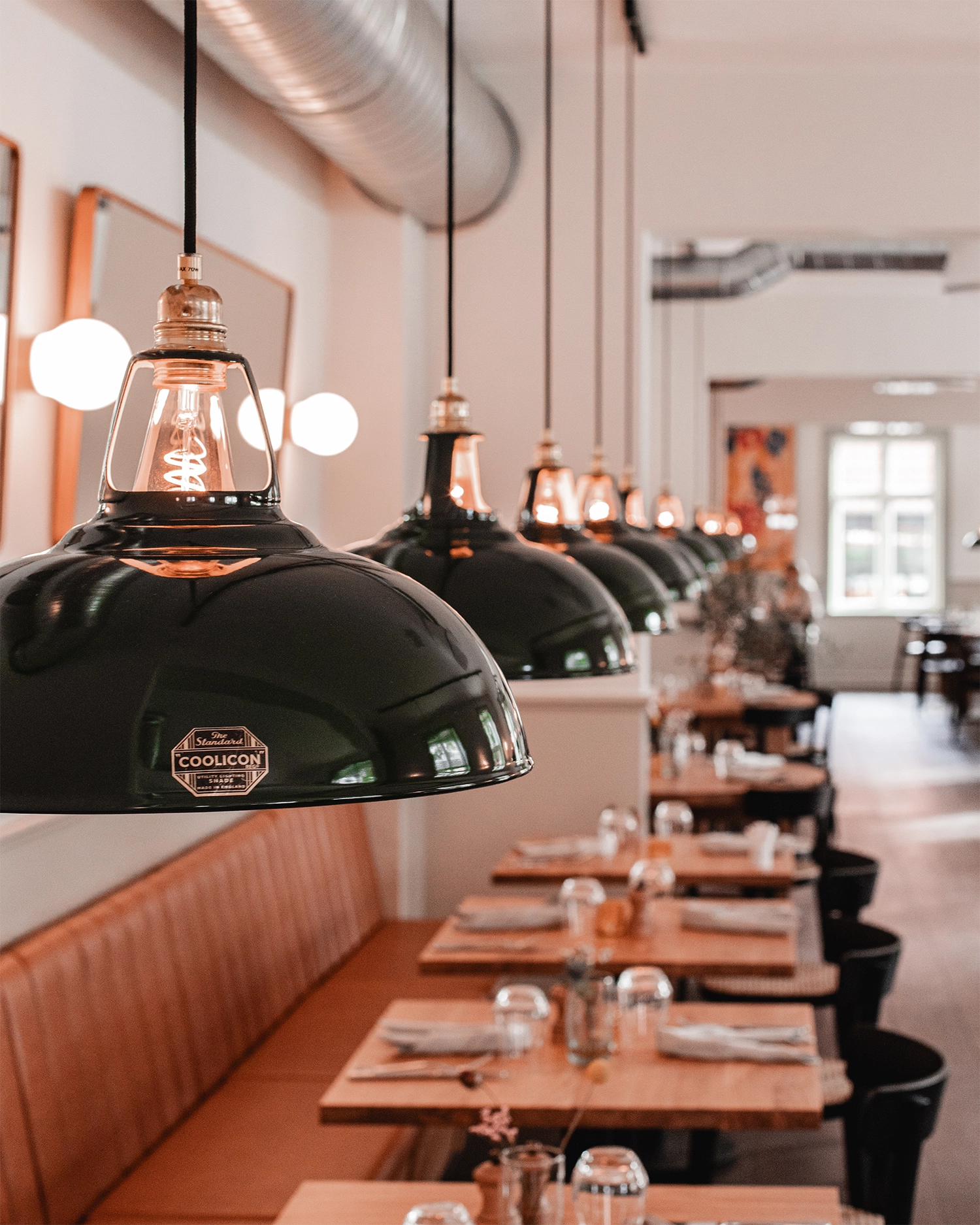 A close up of a neat row of evenly spaced Large green Coolicon Lampshades hanging over wooden restaurant tables.