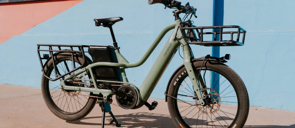 a green Momentum PakYak E+ electric cargo bike in front of a blue wall