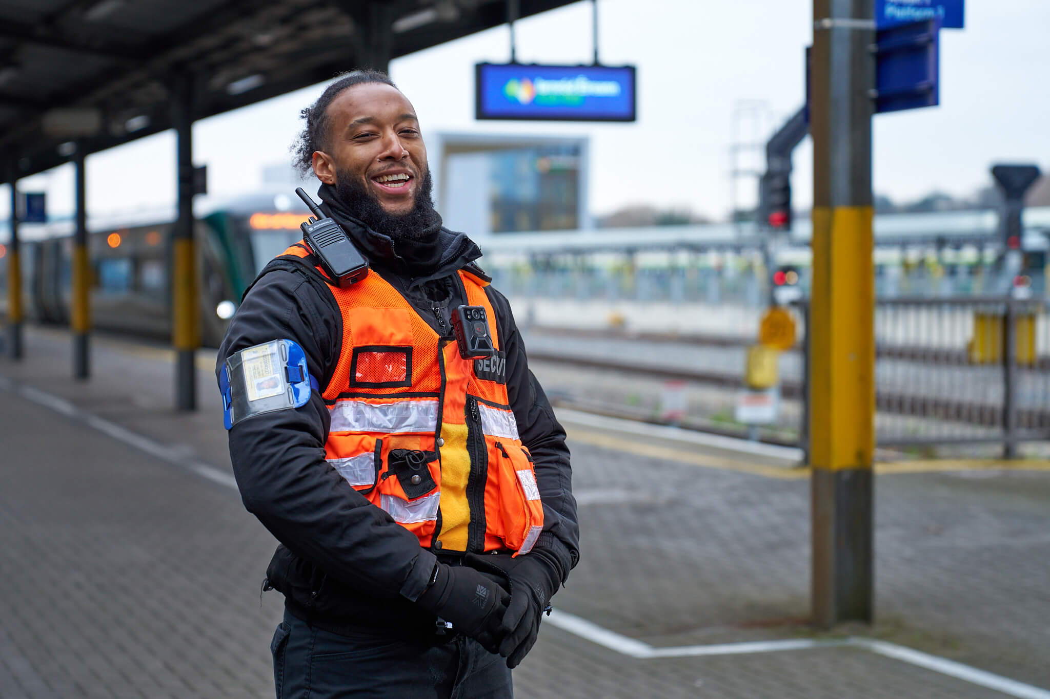 A person wearing an orange safety vest and black gloves stands smiling on a train station platform. The background shows tracks, a train, and signage.