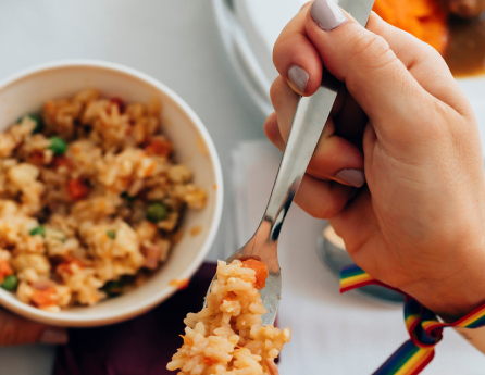 A person holding a fork with rice over a bowl of rice mixed with vegetables. They are wearing a rainbow-colored wristband on their right wrist.