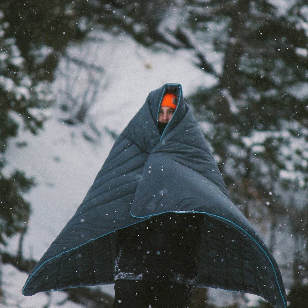 Man bundled up standing in the snow wrapped in a Rumpl camping blanket