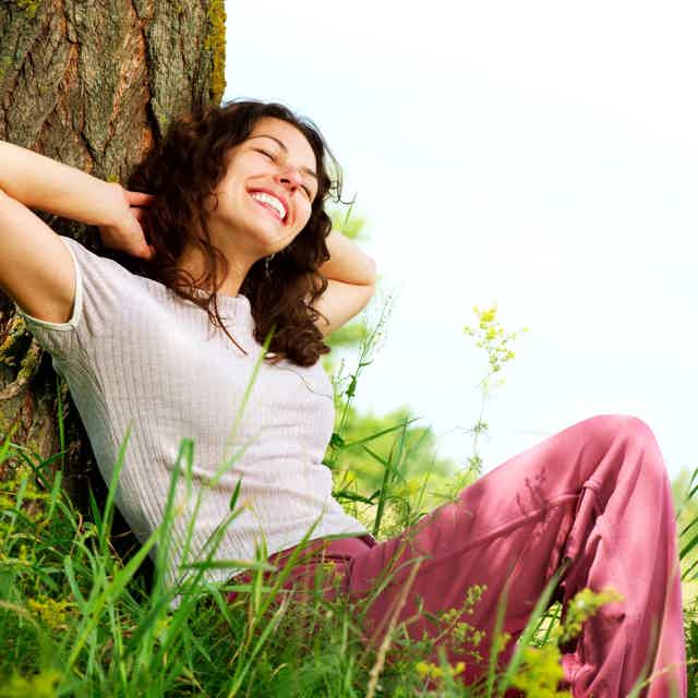 Woman relaxing against a tree in a sunny field, smiling with hands behind head and pink pants.