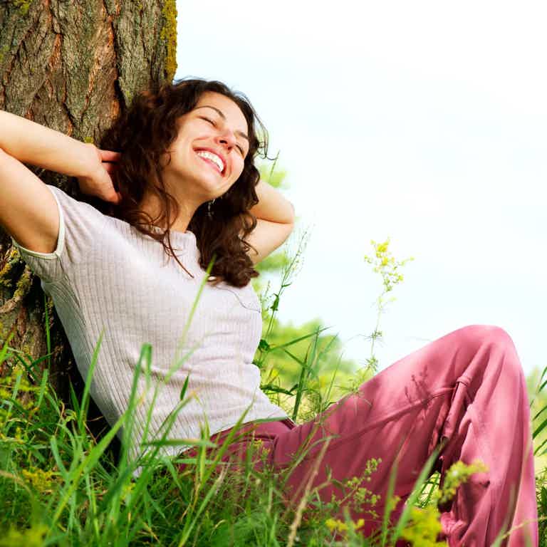 Woman relaxing against a tree in a sunny field, smiling with hands behind head and pink pants.