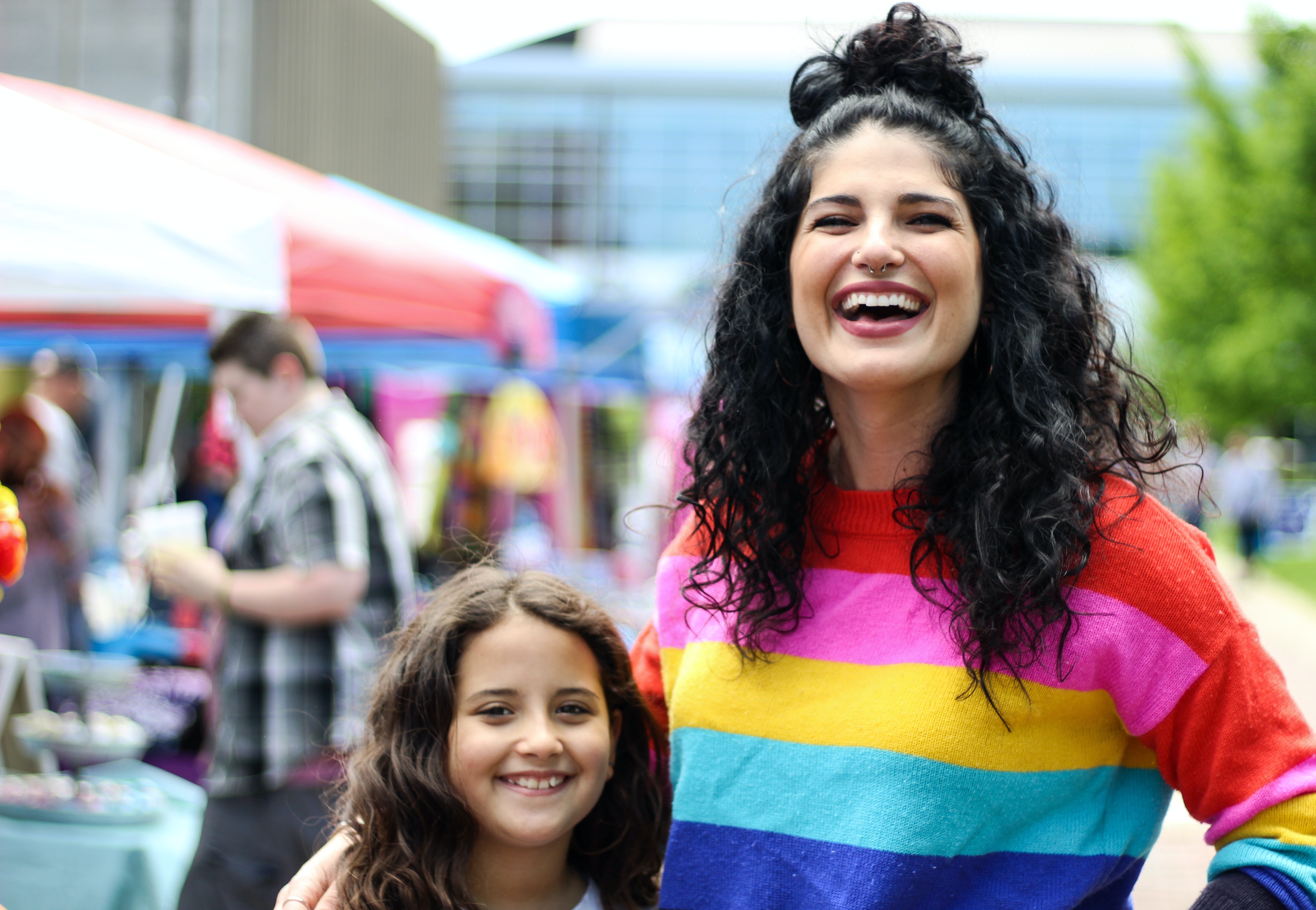 A woman wearing a rainbow sweater laughs while standing next to her young daughter smiling as well.