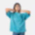 A woman wearing a loose teal top with wide sleeves, arms raised behind her neck, paired with jeans on a white background, showcasing a relaxed casual style.