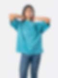 A woman wearing a loose teal top with wide sleeves, arms raised behind her neck, paired with jeans on a white background, showcasing a relaxed casual style.