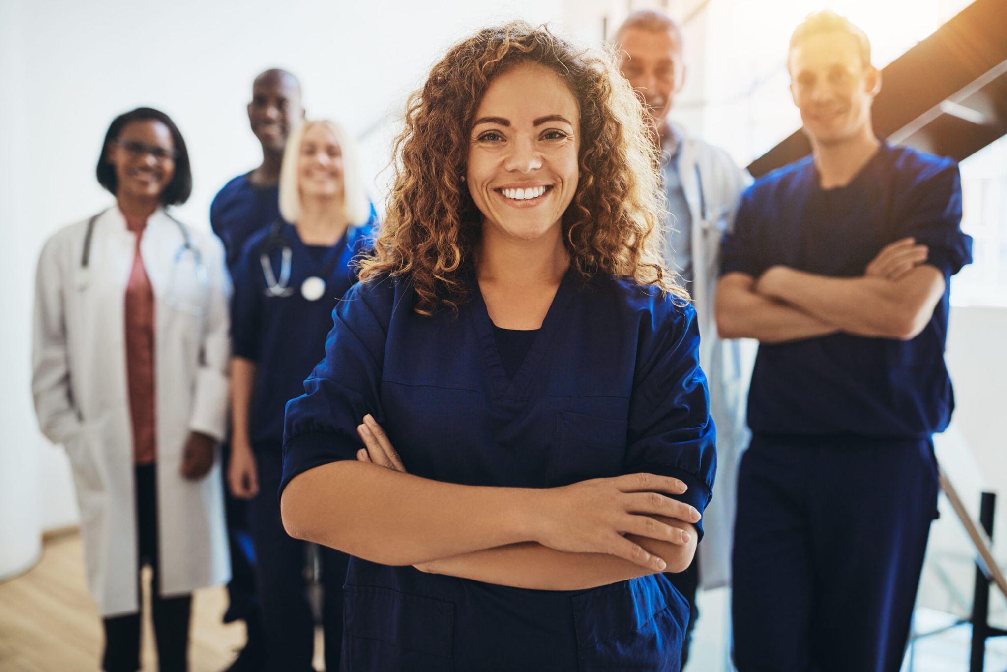 A smiling woman in navy blue scrubs stands confidently with arms crossed in the foreground, while a diverse group of healthcare professionals in scrubs and lab coats stand behind her, smiling.
