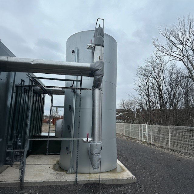 A large vertical metal storage tank next to a building and pipes on a paved outdoor area, with bare trees and a cloudy sky in the background.