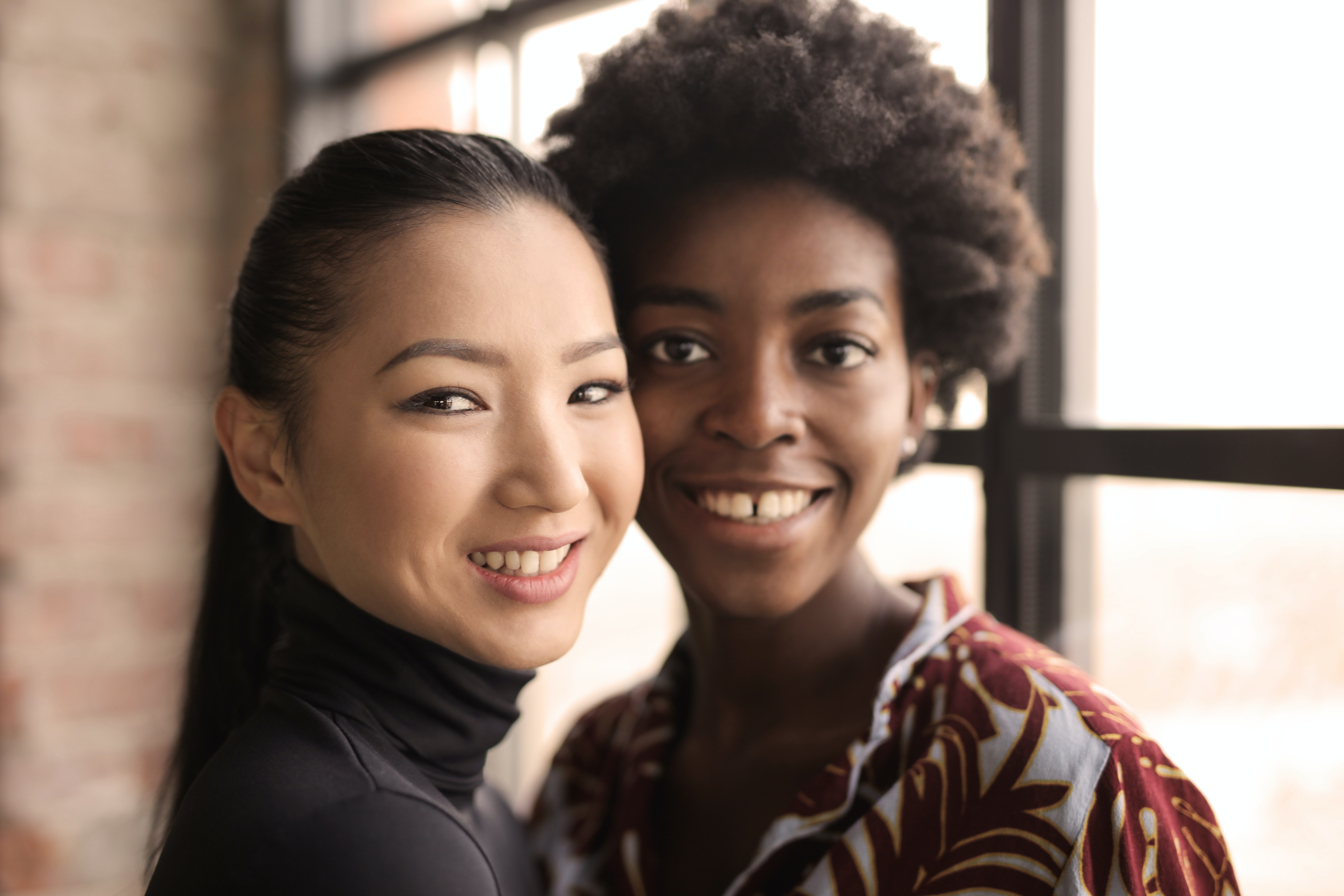 Two attractive black and asian women stand close to eachother pressing their cheeks and smiling to the camera.
