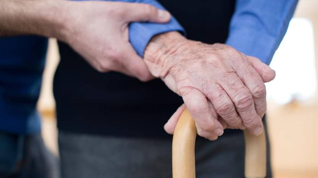 Senior Man's Hands On Walking Stick With Care Worker In Backgrou