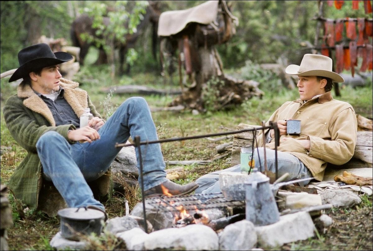 Jack and Ennis from Brokeback Mountain sitting at a campfire talking with a cup in their hands relaxed.