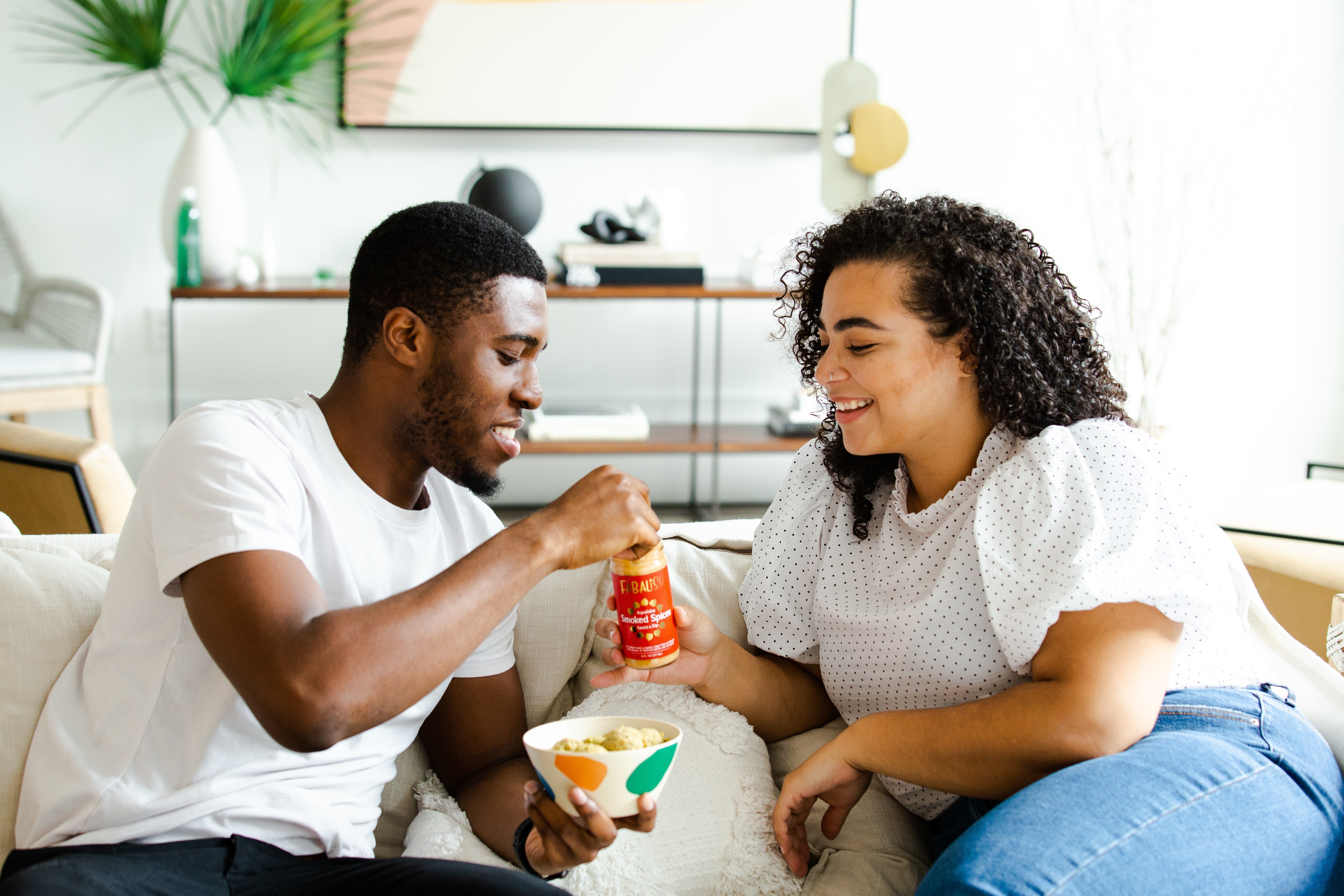 Image of two attractive ethnically diverse man and woman with white t-shirts are sharing snacks on top of a white couch in a sunny living room.