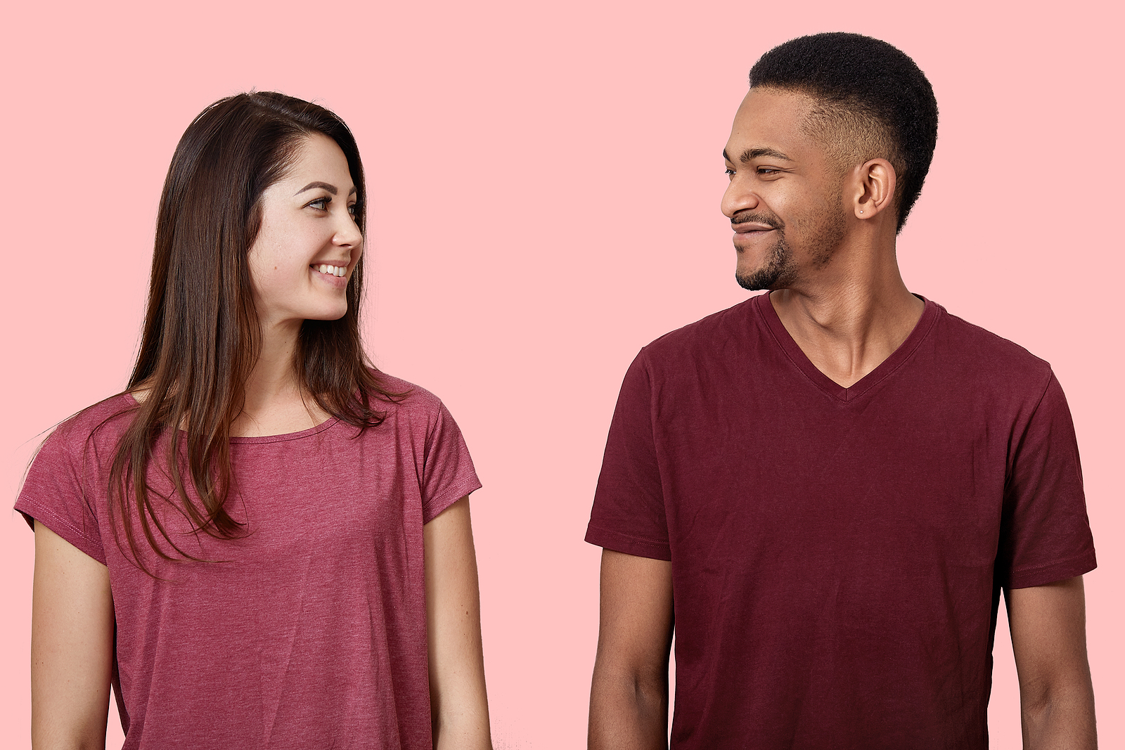 An attractive man and woman smile while standing next to eachother looking at one another against a pink background.
