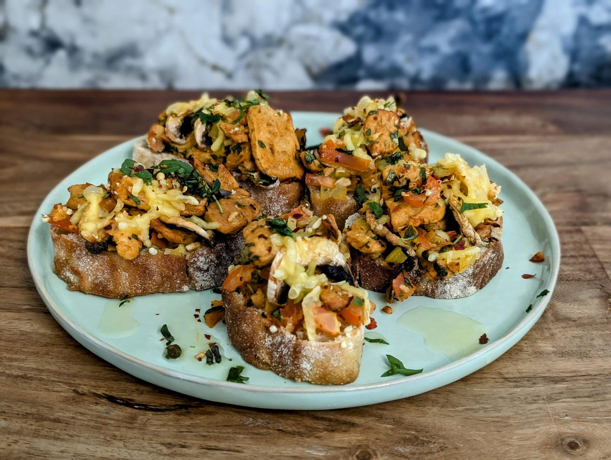 A plate of toasted bread slices topped with a mix of sautéed mushrooms, diced tomatoes, herbs, and melted cheese. The wooden table and a marble-like background add a rustic touch to the presentation.