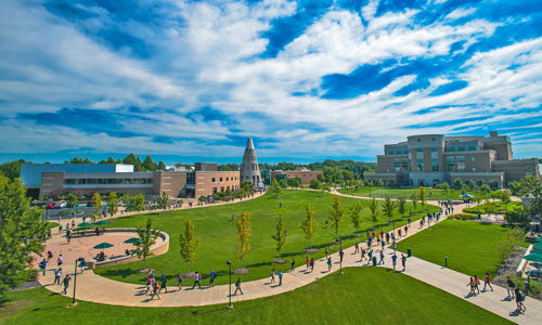 Vibrant campus scene with green lawns, paths, and modern buildings, filled with people, evoking energy and community.