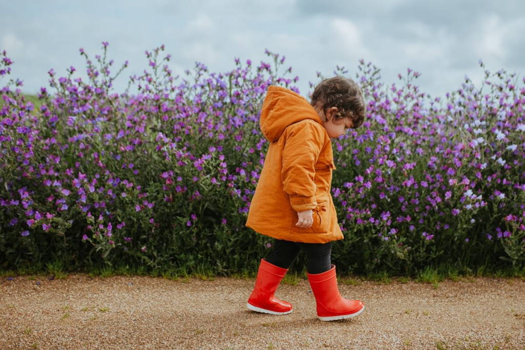 Jeune enfant dans un manteau orange et des bottes de pluie rouges marchant le long d'un chemin à côté d'un champ vibrant de fleurs violettes par une journée nuageuse.