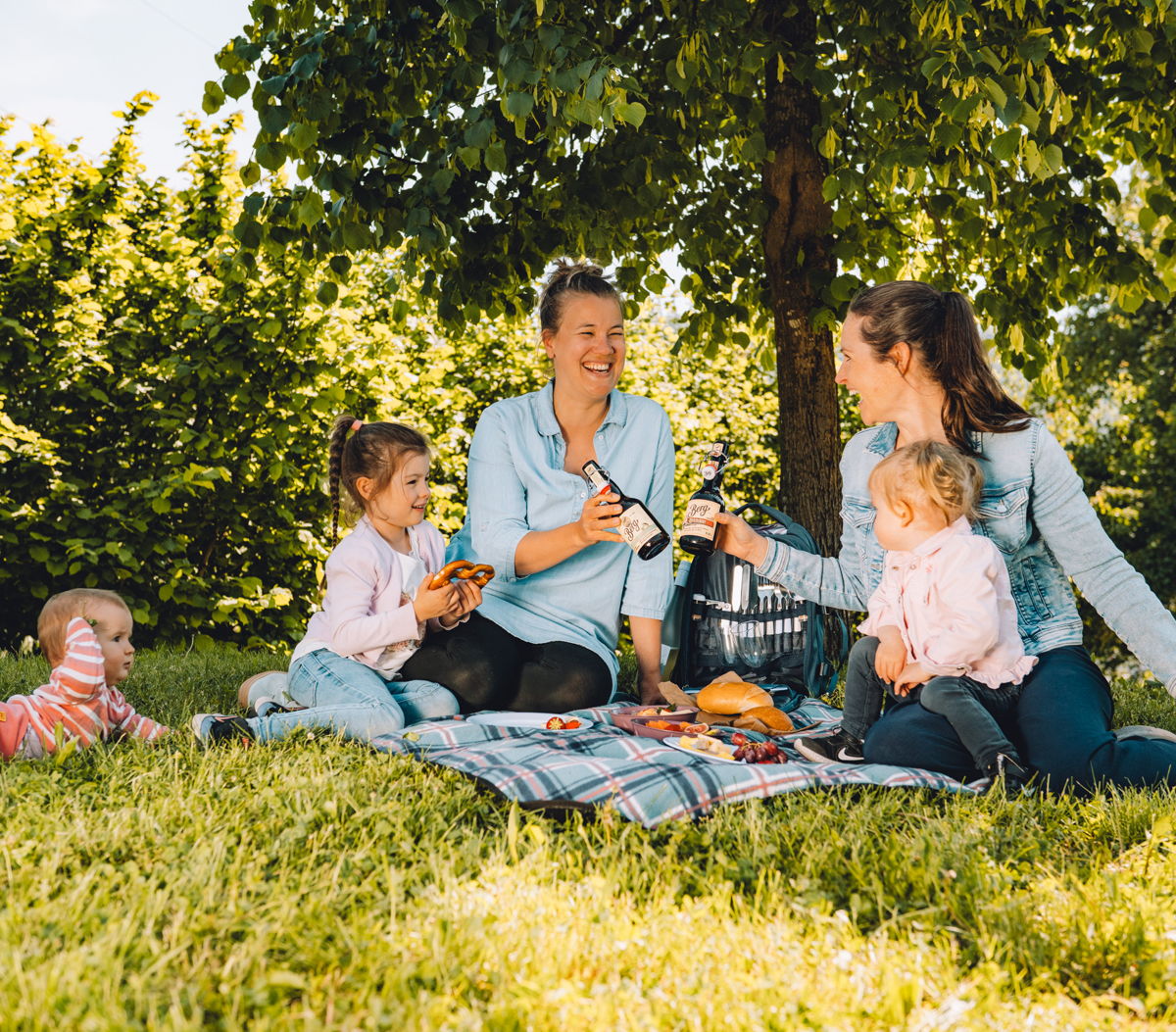 zwei Mütter sitzen mit ihren Kindern auf der Picknick-Decke und stoßen mit einer Flasche an