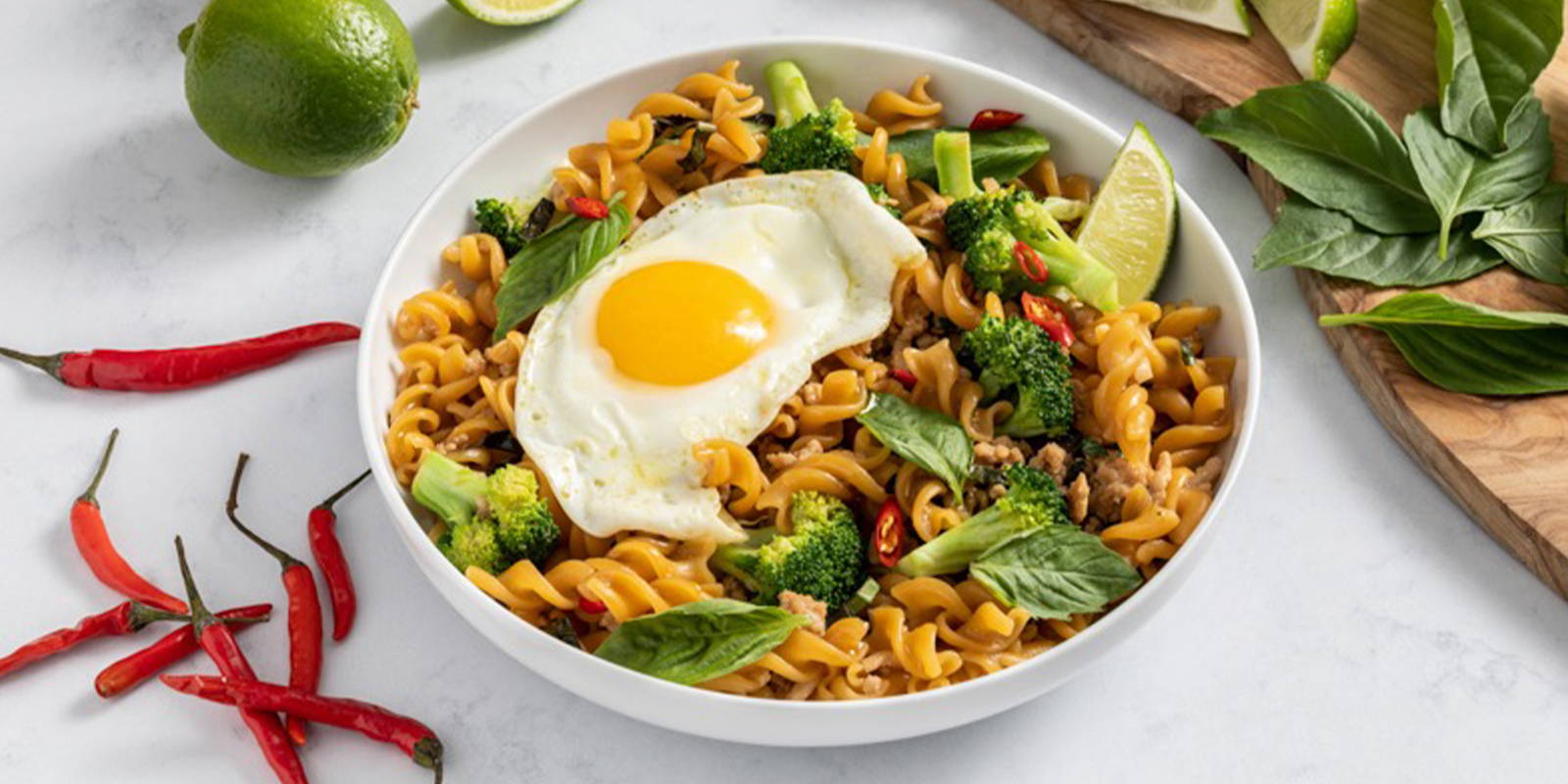 A white bowl of ZENB Thai Style Rotini placed on a white table and styled with limes, chili peppers, and Thai basil.