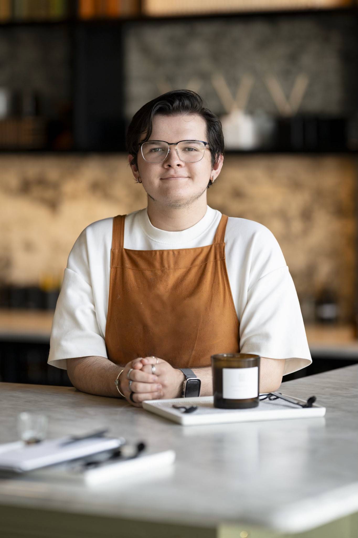 man with glasses in orange apron standing at counter in front of candle bar