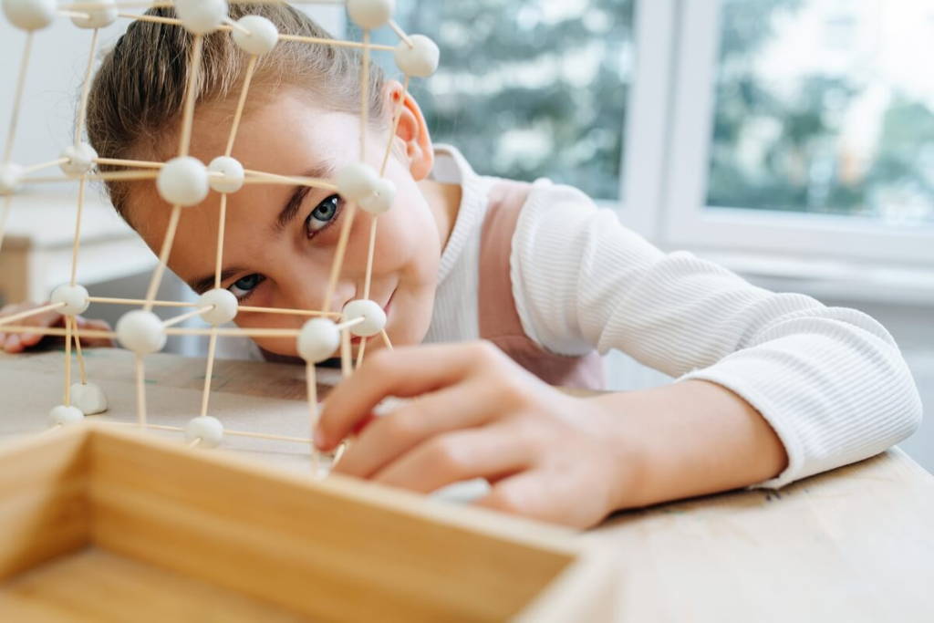 Little girl playing with a DIY structure made out of cotton balls and wooden sticks.