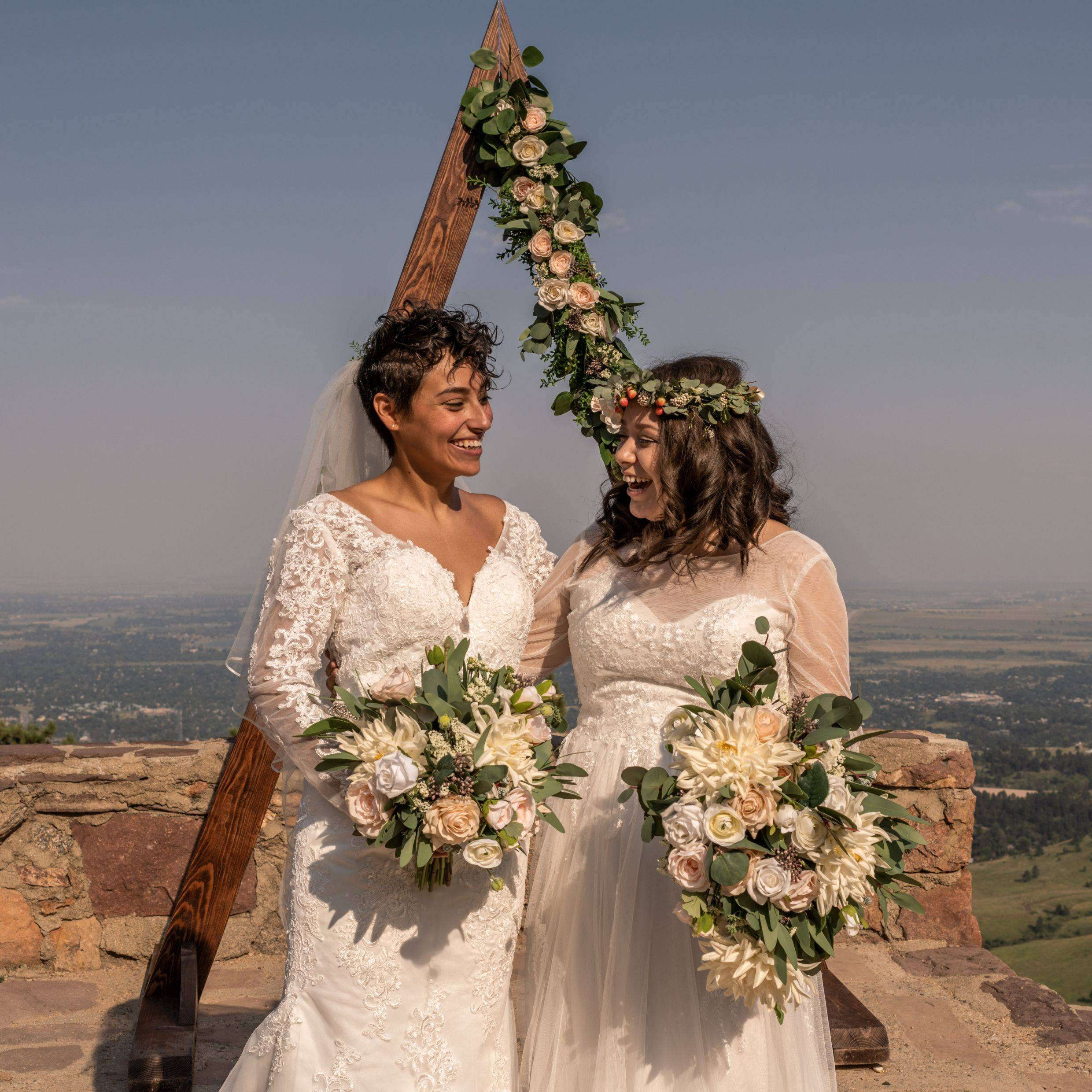 Two brides hold each other and are laughing in front of Compass Rose Floral's wooden triangle arch. They are each holding a bouquet made of cream, blush, and white flowers. The arch has these flowers on it as well 