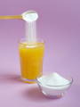 a spoonful of collagen powder being added to a tall glass of orange juice, next to a small dish of multi-collagen protein powder on a lavender purple background