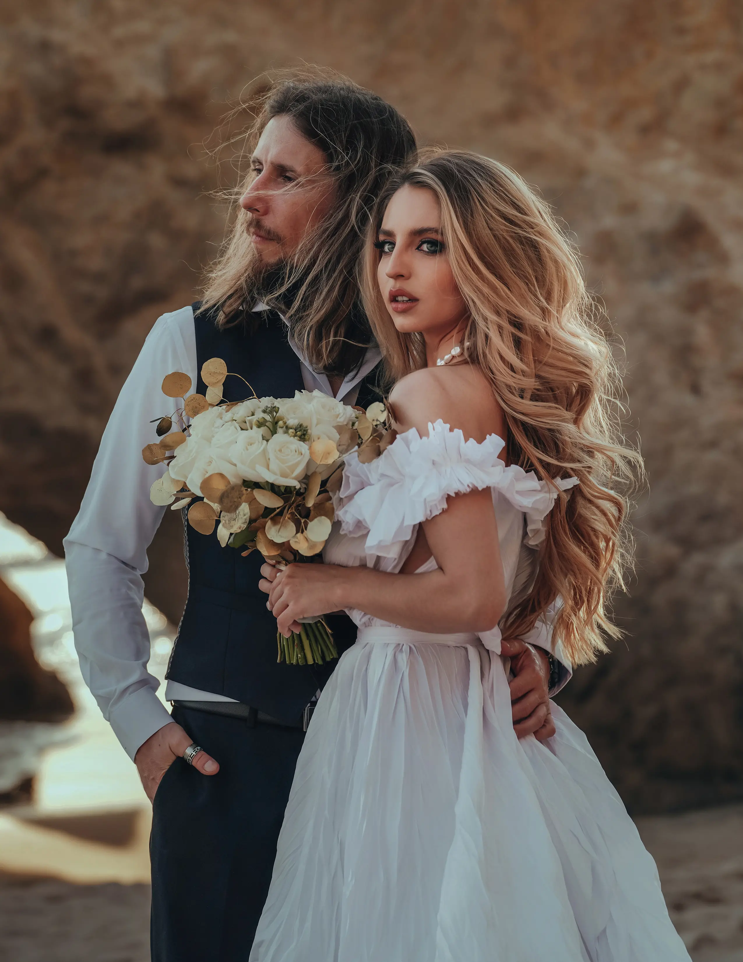 Photo d'une mariée blonde aux cheveux longs dans une robe de mariée bohémienne, tenant un bouquet, avec son mari brun aux cheveux ondulés mi-longs portant un gilet bleu marine sur une chemise noire et un pantalon.