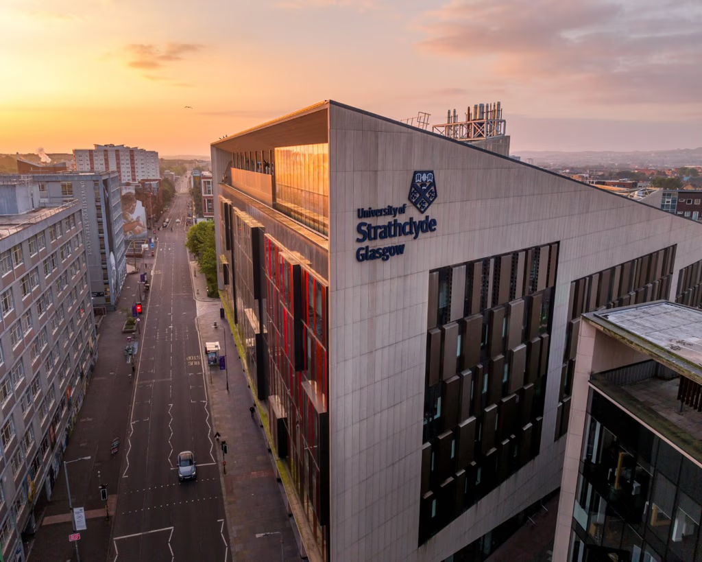 A modern building of the University of Strathclyde in Glasgow at sunset, with quiet streets and cityscape visible in the background. The university’s name and crest are displayed on the building’s facade.