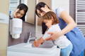 A little girl washing her hands in the sink while her mother holds her.