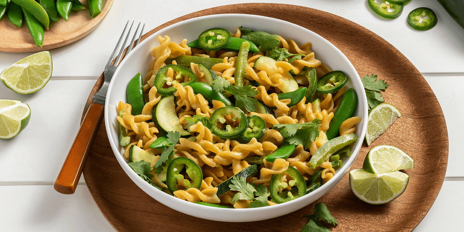 A white bowl of prepared ZENB Green Coconut Curry Rotini atop a wooden plate styled with snap peas, cilantro, lime, and jalapeños.