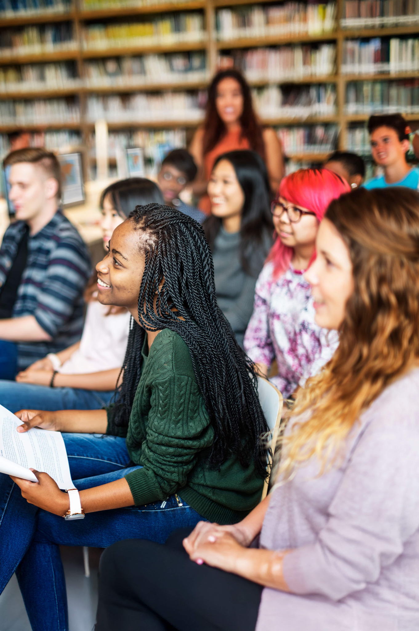 A diverse group of students sitting in a classroom or library, listening attentively and smiling. Bookshelves are visible in the background, and one person holds a paper in their hands.