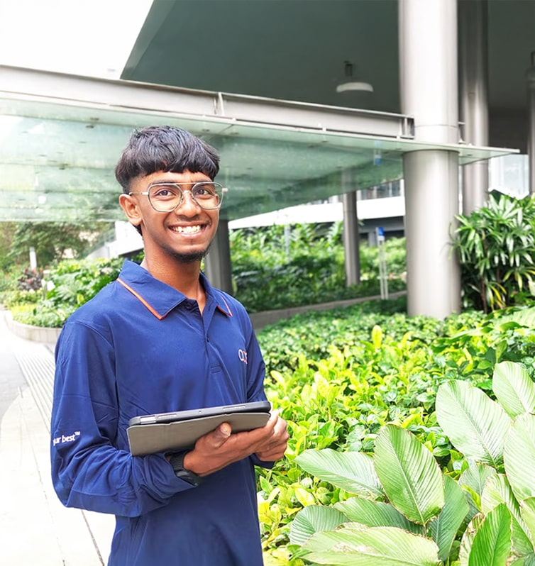 A young man wearing glasses and a blue shirt stands outdoors by lush green plants, smiling and holding a tablet. Modern buildings with glass and metal elements are visible in the background.