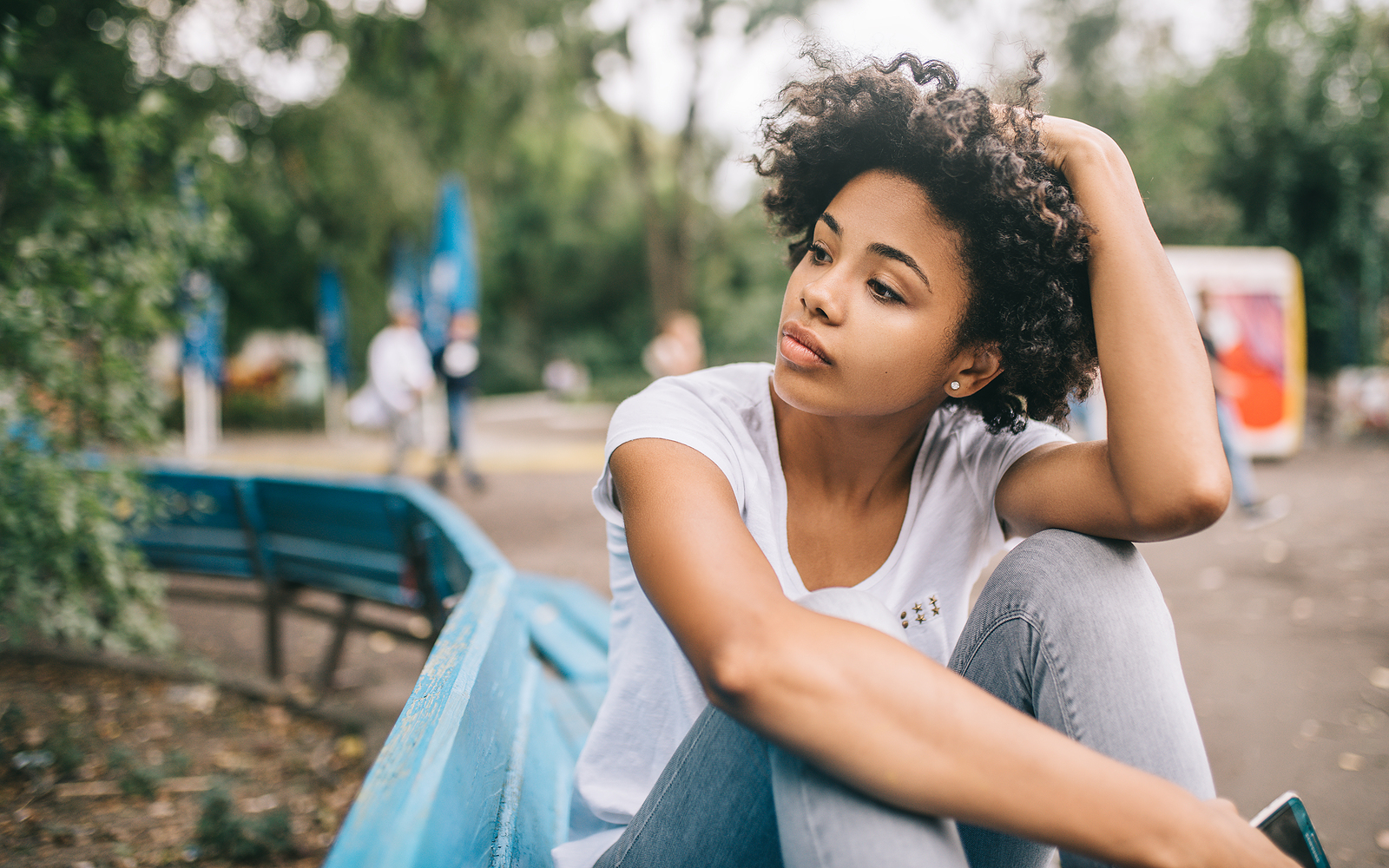 A young black woman with curly hair sits alone on a bench looking to her side contemplative.
