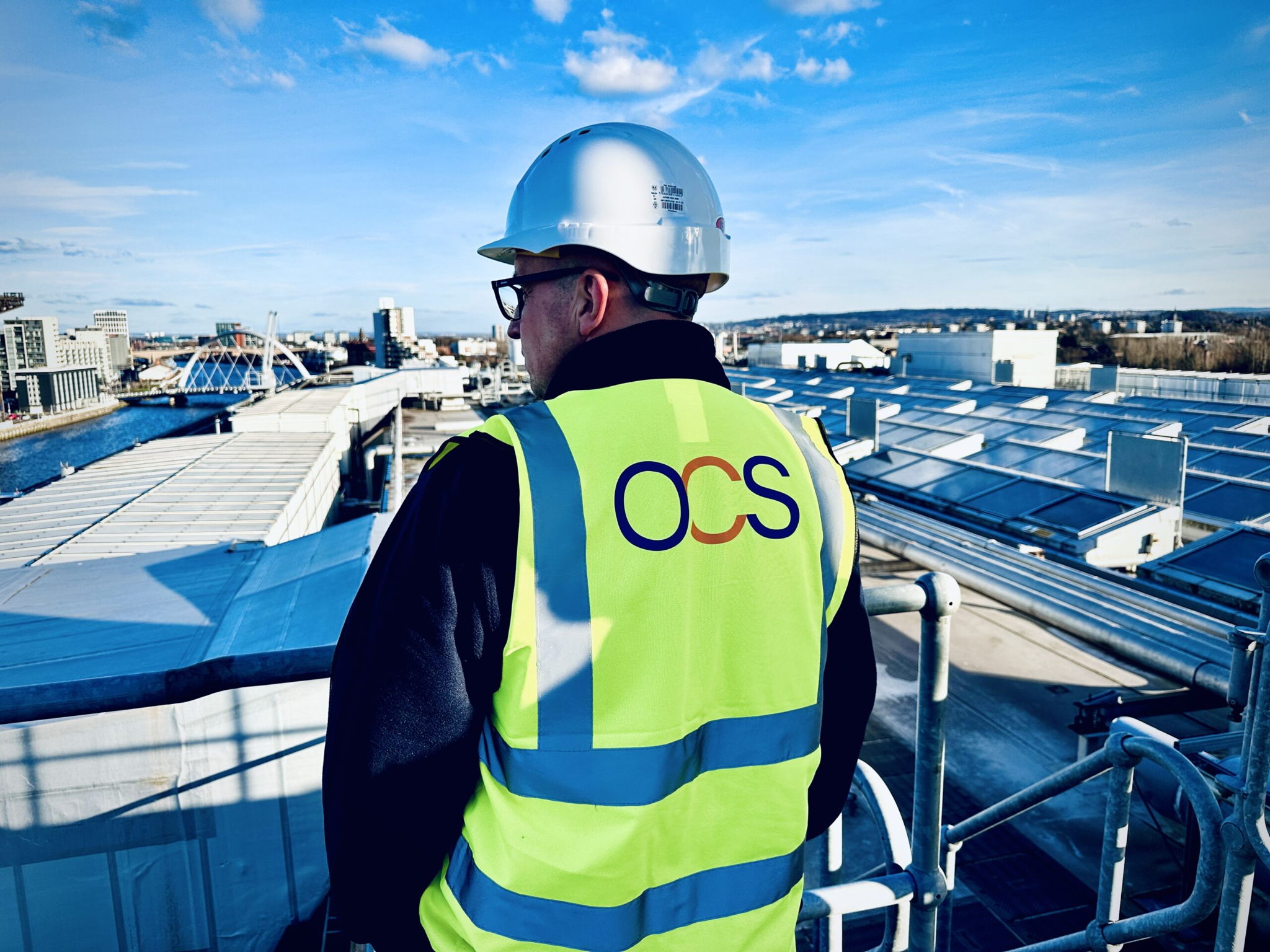 A person wearing a white hard hat and a yellow high-visibility vest with OCS on the back stands on a rooftop overlooking an industrial area and a river under a blue sky.
