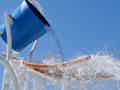 Ein großer blauer Eimer kippt Wasser auf einen Wasserspielplatz unter einem klaren blauen Himmel.