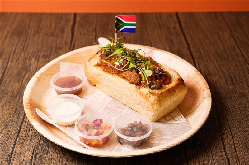A hollowed bread loaf filled with curry, topped with herbs and a small South African flag, served on a wooden plate with four small containers of sauces and salsa on the side.