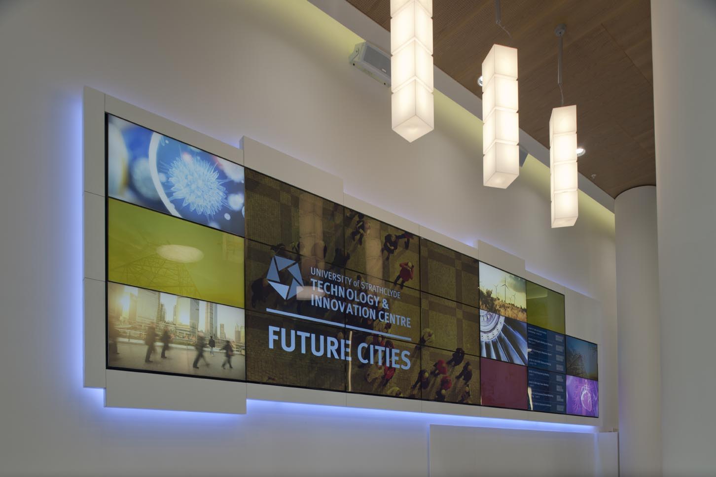A modern video wall displays University of Strathclyde Technology & Innovation Centre FUTURE CITIES with various colorful images; contemporary pendant lights hang from the ceiling above.
