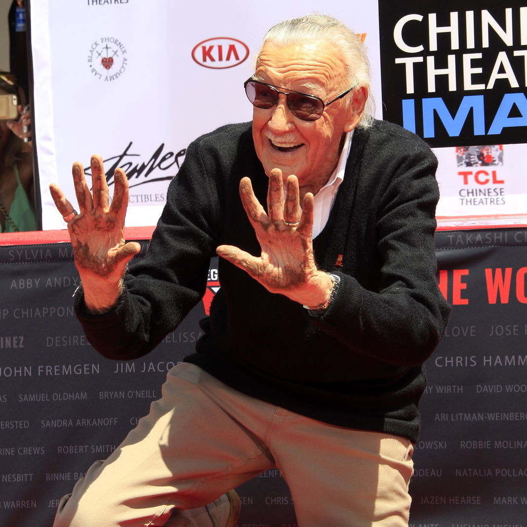 Stan Lee smiling at a carpet event, showing his hands covered in concrete after making a stamp.