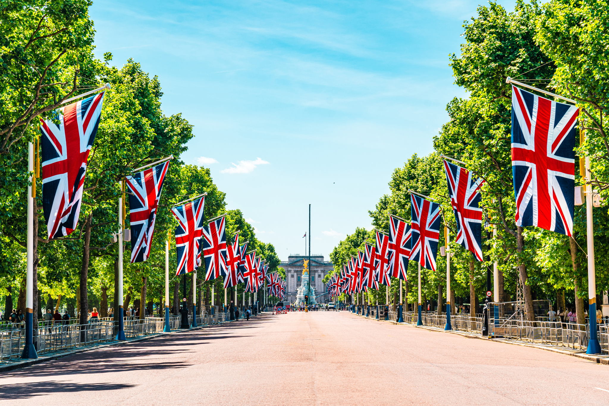 A wide road lined with tall green trees and numerous Union Jack flags, leading towards a historic building in the distance under a bright blue sky. People stand behind barriers along the sides of the road.